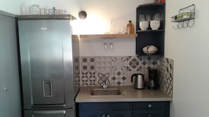 Een compacte keuken met een koelkast, een spoelbak en een koffiezetapparaat. Boven de spoelbak zijn er twee plankjes, waarvan er één beker glazen bevat en de ander een kom.