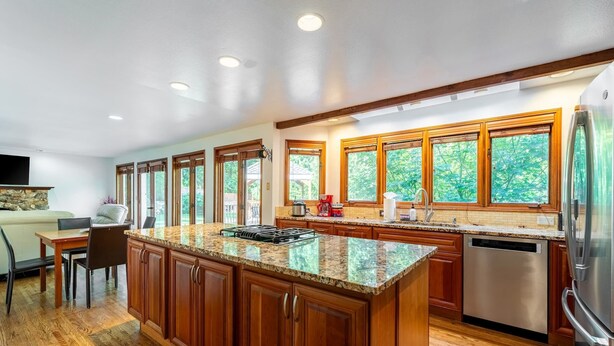 A modern kitchen with wooden cabinets, granite countertops, a built-in island, stainless steel appliances, and large windows offering a view of greenery. - Spacious 3BR Redmond Home with Fireplace & BBQ (Redmond)