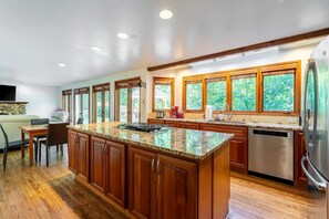 A modern kitchen with wooden cabinets, granite countertops, a built-in island, stainless steel appliances, and large windows offering a view of greenery. - Spacious 3BR Redmond Home with Fireplace & BBQ (Redmond)