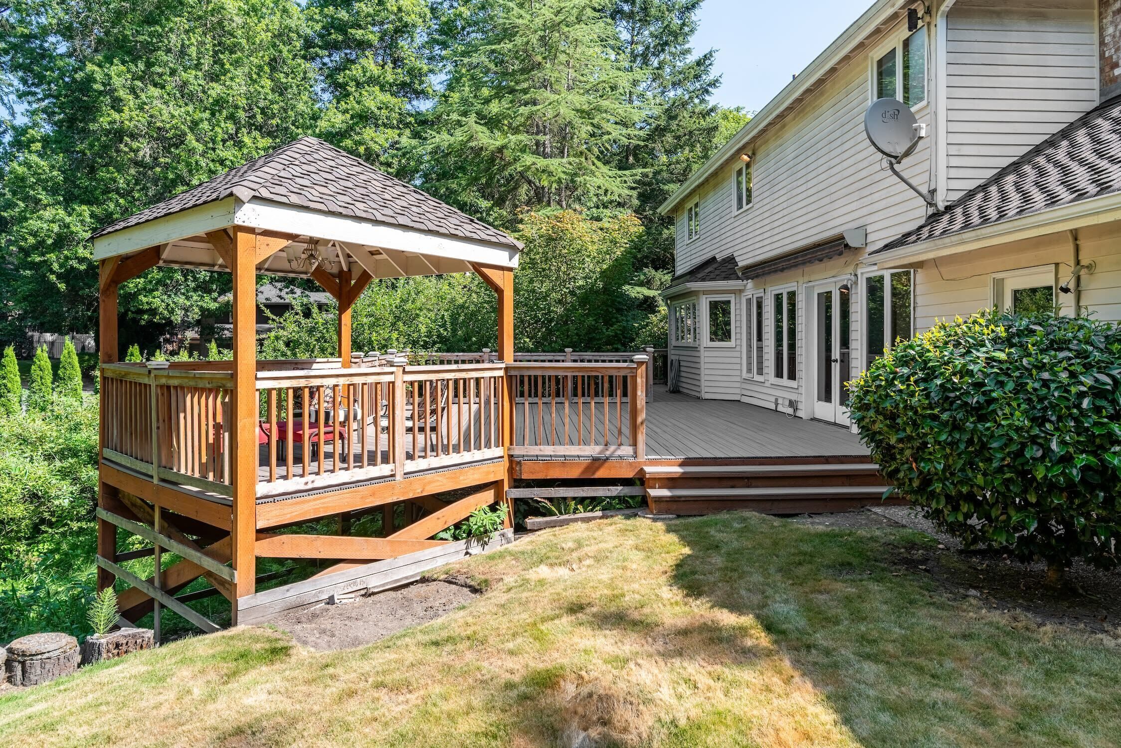 A wooden gazebo with a covered deck, a fenced deck area, and a house with a satellite dish.