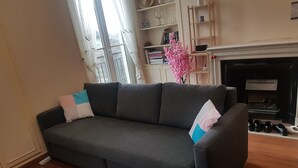 A dark grey sofa with two pillows, a wooden floor, a bookshelf with books and decorative items, and a fireplace.