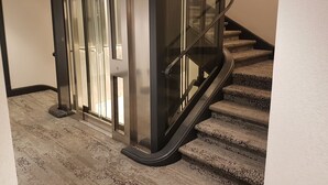 A modern elevator with glass doors and a metal frame, next to a carpeted staircase with a handrail.