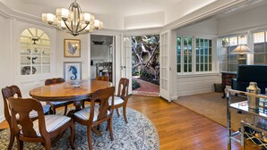 A dining area with a round wooden table, six chairs, and a chandelier. There is a view of a garden through an open door. - Oceanview Charmer, One Block to Beach (San Diego)