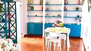 A spacious room with a wooden floor, a white dining table with chairs, a blue shelving unit with plants, and a green pendant light. - Delightful and spacious apartment with a view of Vesuvius - Salotto Napoletano 381 (Napoli)