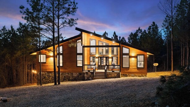 Une cabane en bois à deux étages, avec des fondations en pierre, de grandes fenêtres et une allée gravillonnée.