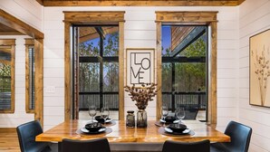 Un coin repas avec une table en bois, des chaises noires et une vue sur les arbres à travers de grandes fenêtres.