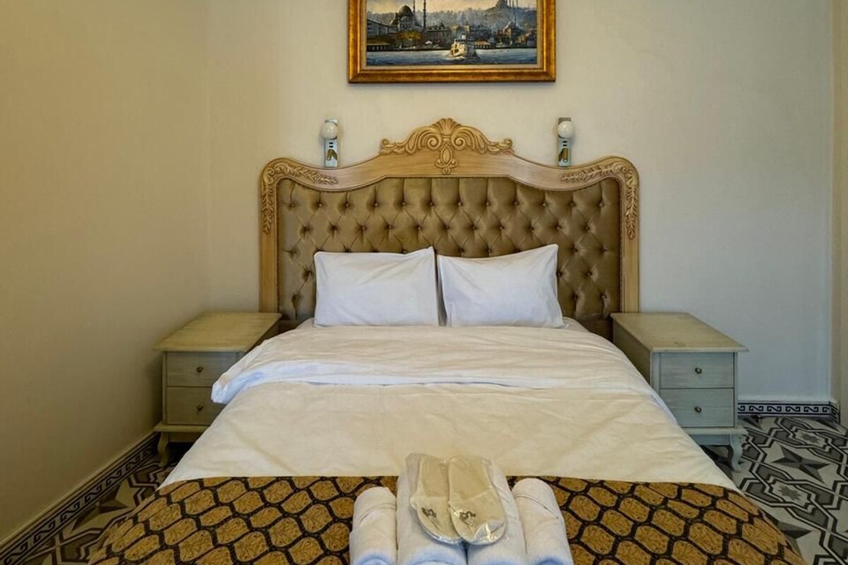 A hotel room with a tufted headboard, a bed with white linens, a painting of a cityscape, and a patterned floor.