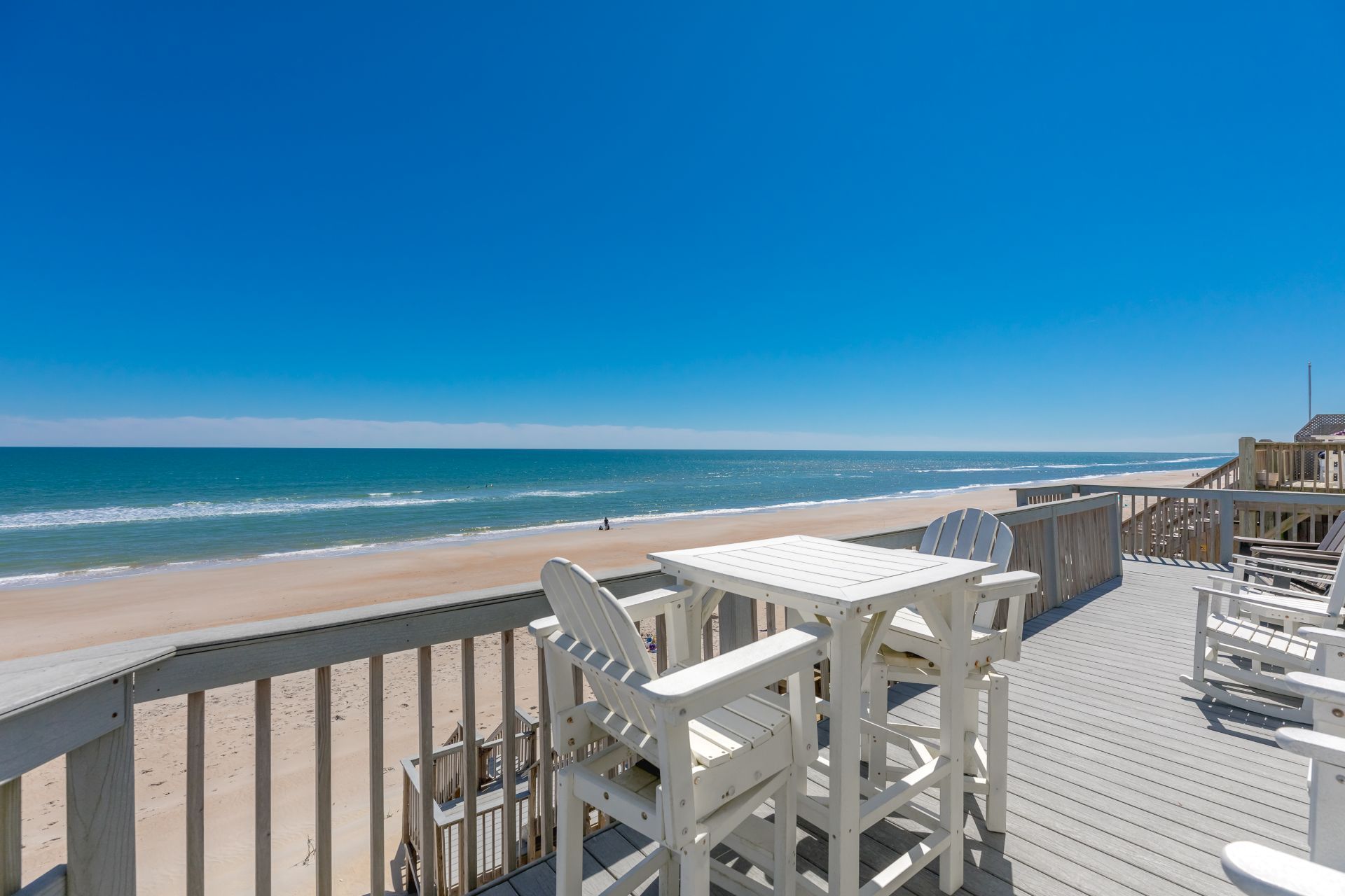 Una terraza de madera con sillas y mesas blancas con vista a la playa.