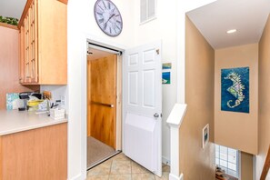 Un pasillo con una puerta de madera, una puerta blanca y un reloj de pared. Hay un cuadro en la pared y una pequeña mesa con objetos encima.