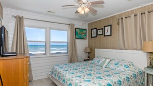 Un dormitorio con cama, escritorio, silla, ventana con cortinas, ventilador de techo y pared con cuadros.