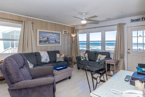 A living room with a view of the ocean, a ceiling fan, and a door labeled 'SANDY'. - Carolina Charm - Classic 3BR Oceanfront Cottage, Dog Friendly! (Surf City)