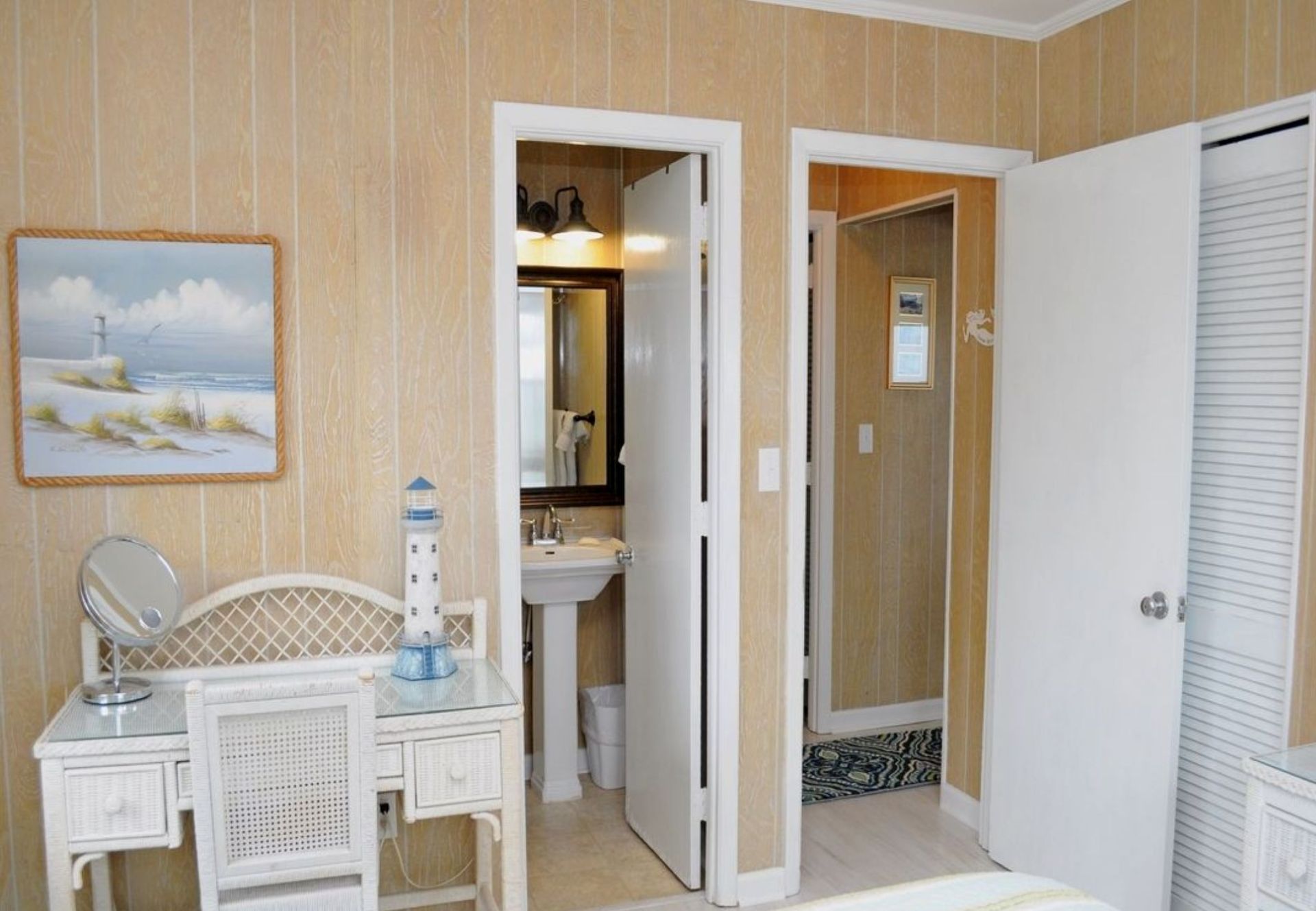 Une chambre d’hôtel comprenant un lit, une coiffeuse avec un miroir, une petite table, une salle de bain avec un lavabo et un miroir, et un tableau représentant un phare.