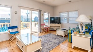 A living room with a TV, a blue chair, a sofa, a lamp, and a glass coffee table. - Almost Beachfront - Cozy 2nd Row Home with 4 Bedrooms (Surf City)