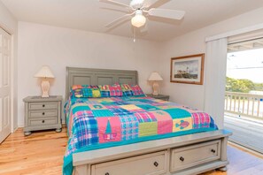 A bedroom with a bed, two bedside tables, a ceiling fan, a window with a view, and a framed picture on the wall. - Almost Beachfront - Cozy 2nd Row Home with 4 Bedrooms (Surf City)