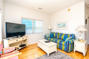 A living room with a flat-screen TV, a striped sofa, a white coffee table, and a small cabinet. - Almost Beachfront - Cozy 2nd Row Home with 4 Bedrooms (Surf City)