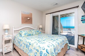 A bedroom with a bed, two bedside tables, a nightstand, a window with a view of the beach, and a door leading to a balcony.