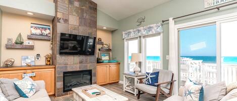 A living room with a stone fireplace, a television, and a view of the ocean through large windows.