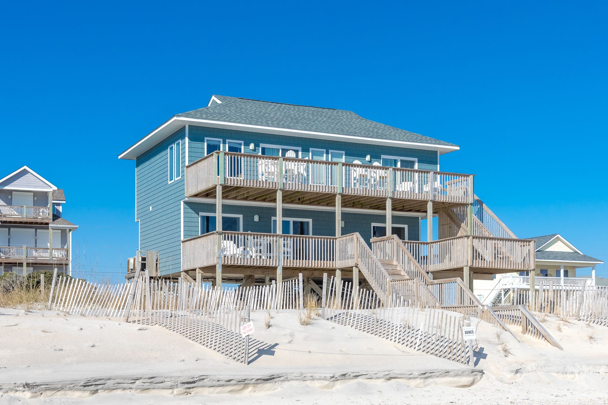 A multi-story beachfront building with balconies, a wooden deck, and a sandy beach in the foreground.