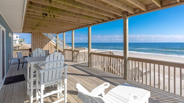 A wooden deck with white chairs and a table, overlooking a beach.