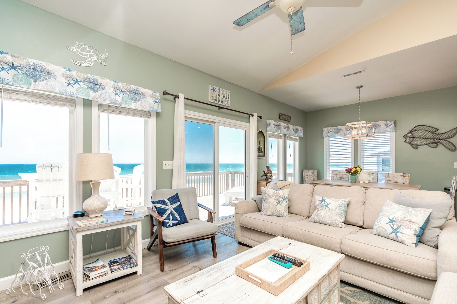 A living room with a view of the ocean, a ceiling fan, and a sofa with star-patterned pillows.