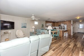 A modern living room with a sofa, a dining area with chairs, a kitchen with wooden cabinets, and a ceiling fan.