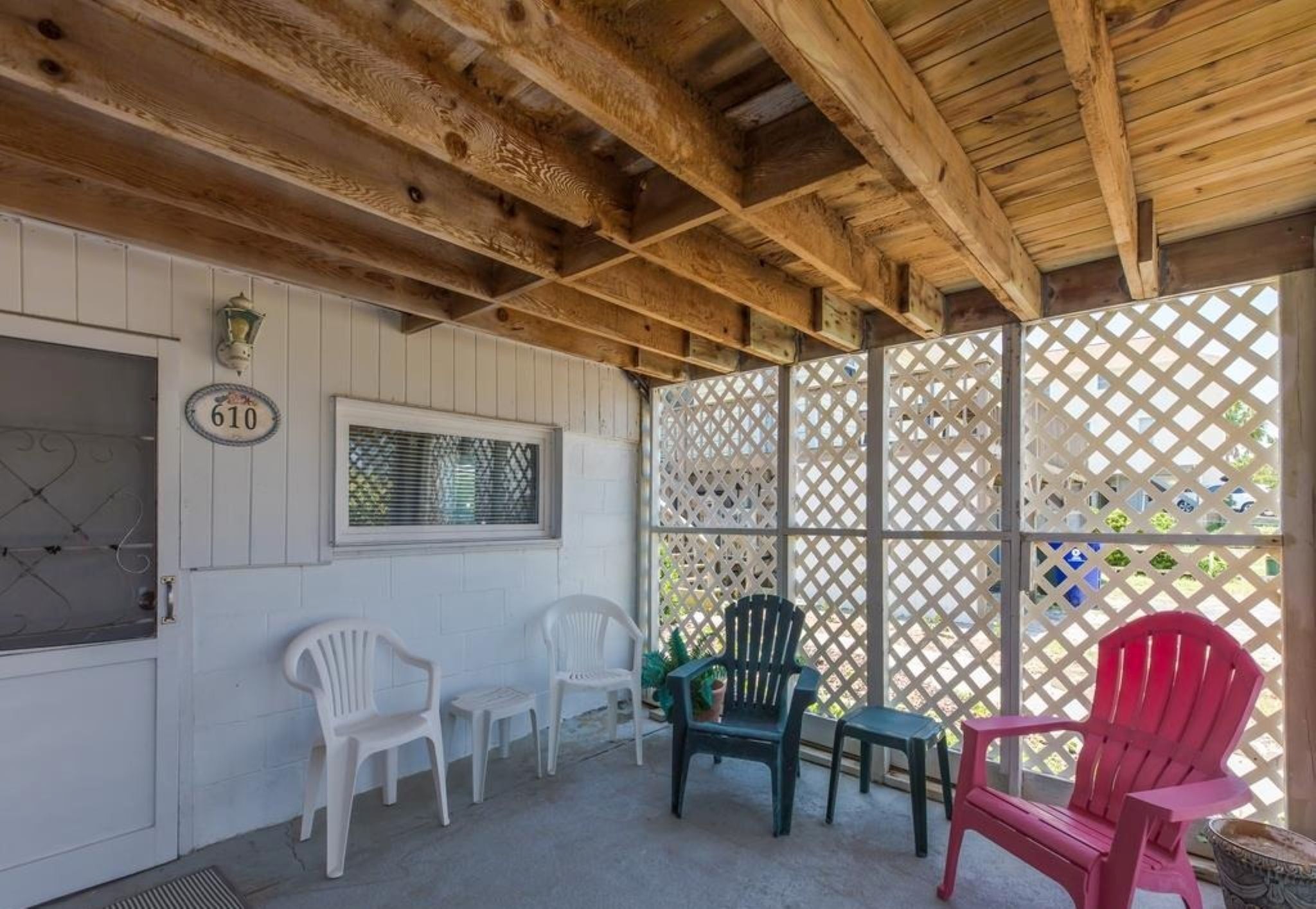 A covered outdoor seating area with wooden beams, a window with blinds, and plastic chairs.