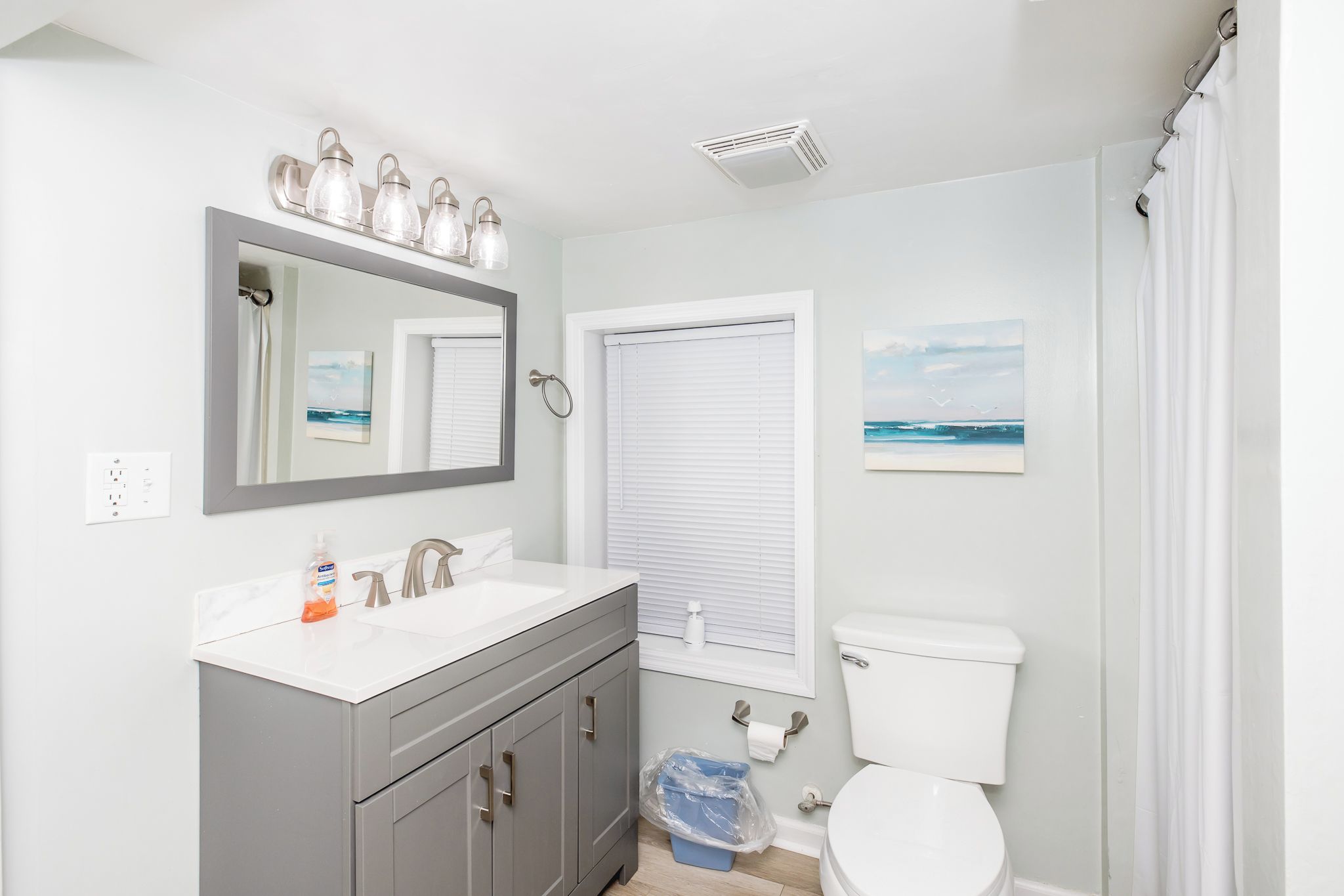 A bathroom with a white sink, mirror, and toilet, a window with blinds, and a shower curtain.