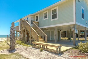 Een strandhuis met een houten trap naar een balkon, een overdekt terras en een zandstrand op de achtergrond.