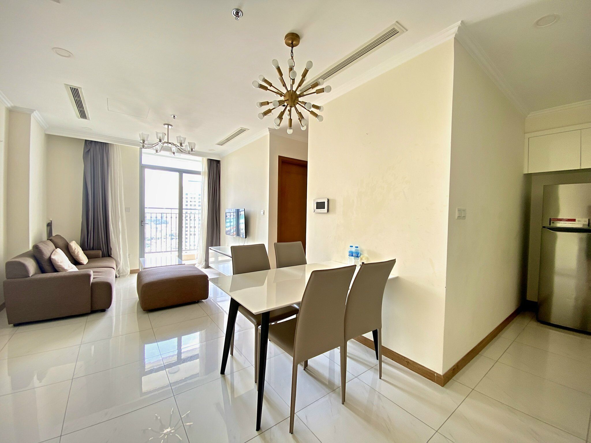 Una habitación de hotel moderna con zona de comedor, sofá, televisión y balcón con cortinas.