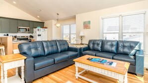 A modern living room with a dark blue leather sectional sofa, a wooden coffee table with magazines, and a kitchen in the background. - Dakota Dunes - Oceanfront!  Dog Friendly! (Surf City)