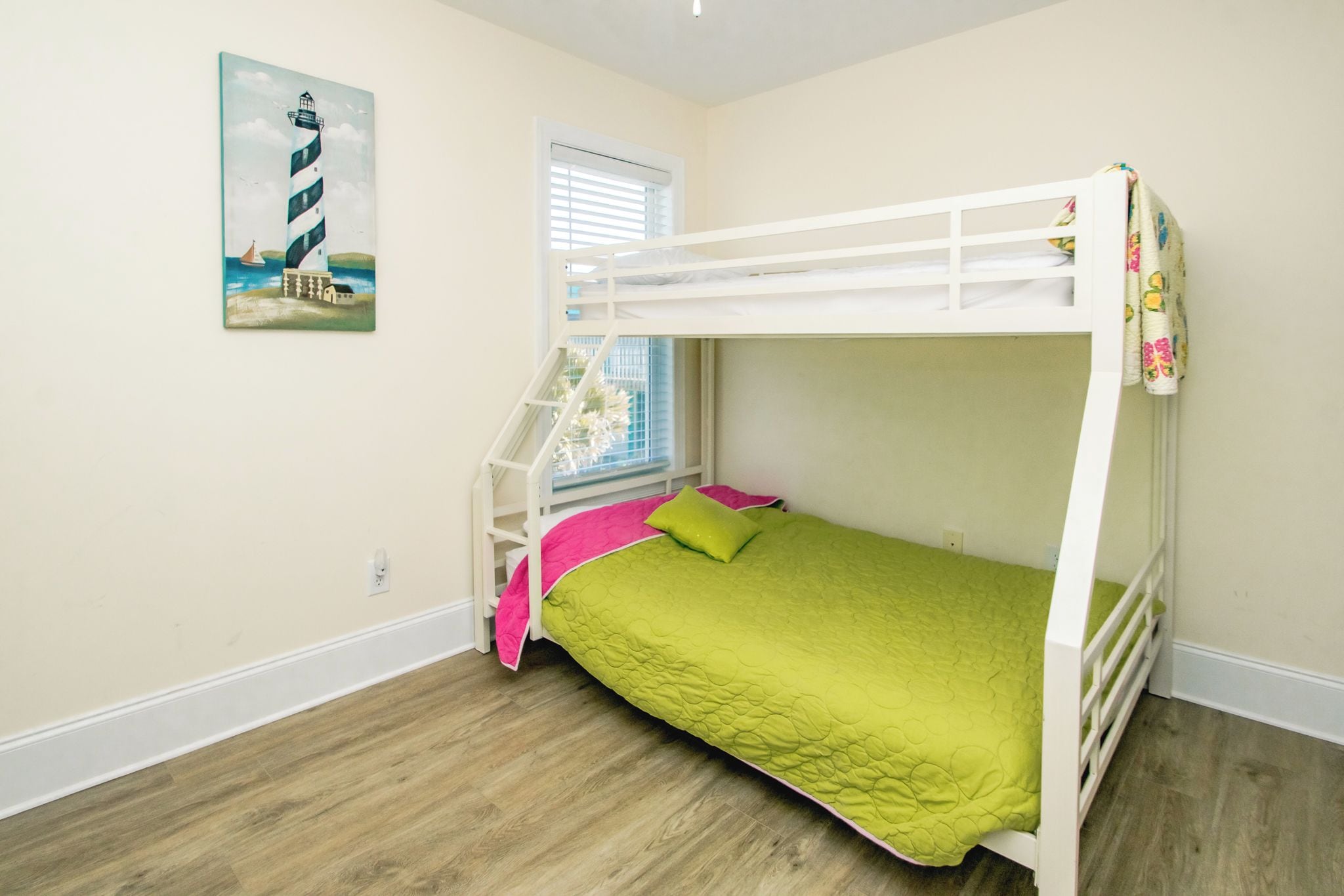 A bunk bed with a green bedspread and pink pillows, a window with blinds, and a painting of a lighthouse on the wall.