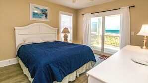 Une chambre à coucher comprenant un lit, une table de chevet, une lampe, un ventilateur de plafond et une vue sur la plage, accessible par une porte coulissante.