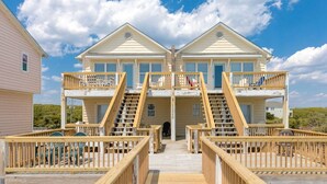 Une maison à deux étages située en bord de mer, dotée d’une terrasse en bois et d’un escalier menant à l’étage supérieur.