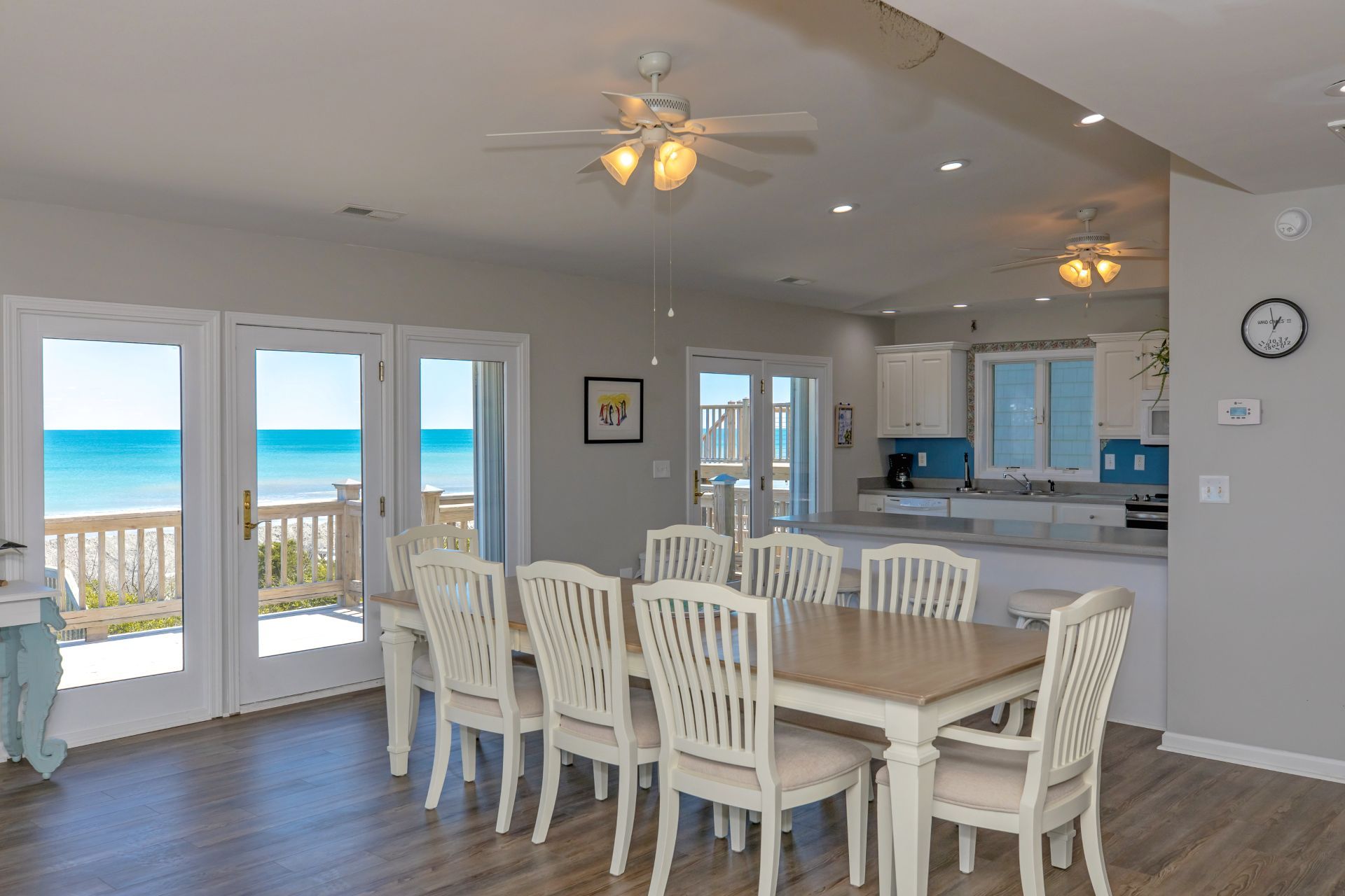 A dining area with a wooden table and white chairs, a kitchen with white cabinets, and a view of the ocean through large glass doors.