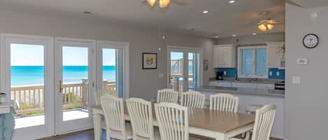 A dining area with a wooden table and white chairs, a kitchen with white cabinets, and a view of the ocean through large glass doors.