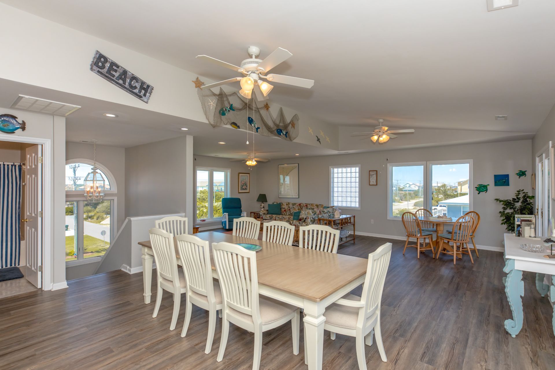 A spacious dining area with a large wooden table and white chairs, a ceiling fan, and a view of the outdoors.