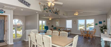 A spacious dining area with a large wooden table and white chairs, a ceiling fan, and a view of the outdoors.