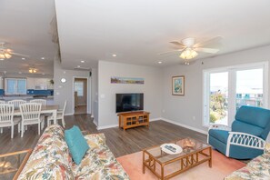 A spacious living room with a television, a dining area with chairs, a sofa with a floral pattern, and a balcony with a view. - Dettor - Oceanfront, Dog Friendly! (Topsail Beach)