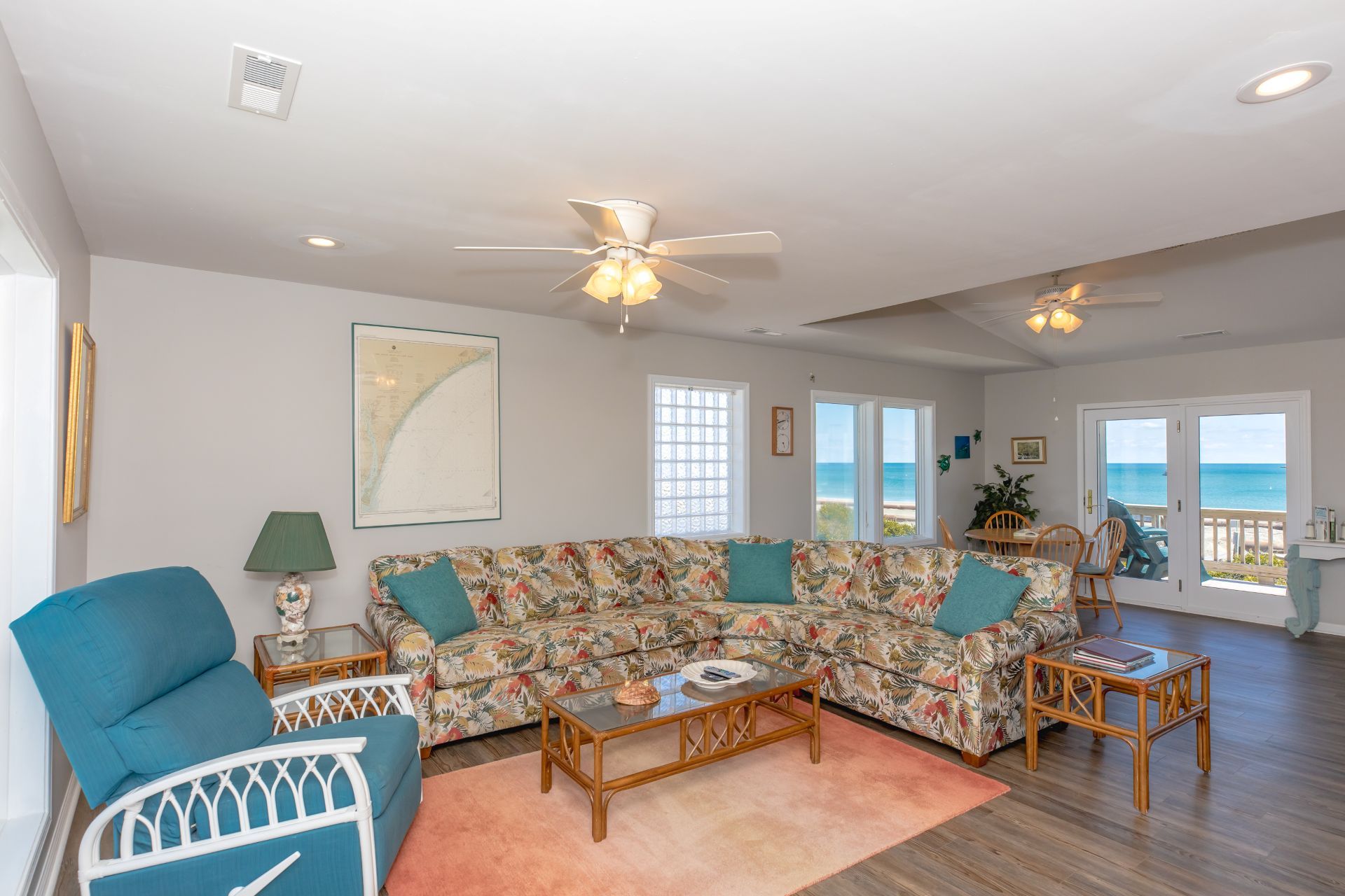 A spacious living room with a large floral sofa, a blue armchair, and a wooden coffee table. There are two ceiling fans, a map on the wall, and a view of the ocean through the glass doors.