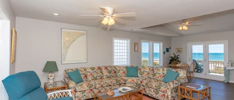 A spacious living room with a large floral sofa, a blue armchair, and a wooden coffee table. There are two ceiling fans, a map on the wall, and a view of the ocean through the glass doors.