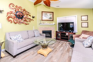 A living room with a sofa, a glass coffee table, a fireplace, a television, and decorative wall art. - Double U Seas - Oceanfront, Dog & Cat Friendly! (Surf City)