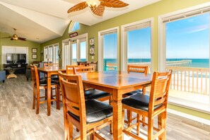 Un comedor con una gran mesa de madera y sillas, un ventilador de techo y vista a la playa.