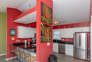 A kitchen with red walls, white cabinets, a stainless steel refrigerator, and a dining area with chairs and a table. - Double U Seas - Oceanfront, Dog & Cat Friendly! (Surf City)