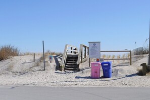 Une zone de plage sablonneuse avec un escalier en bois menant vers le haut, un panneau d’information et deux poubelles.