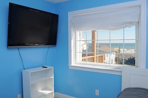 A room with a television mounted on the wall, a window with a view of the beach, and a white shelf unit.