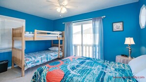 A room with a bunk bed, a single bed, a ceiling fan, a window with curtains, a lamp, and a framed picture on the wall. - Fair Winds - Oceanfront (Surf City)