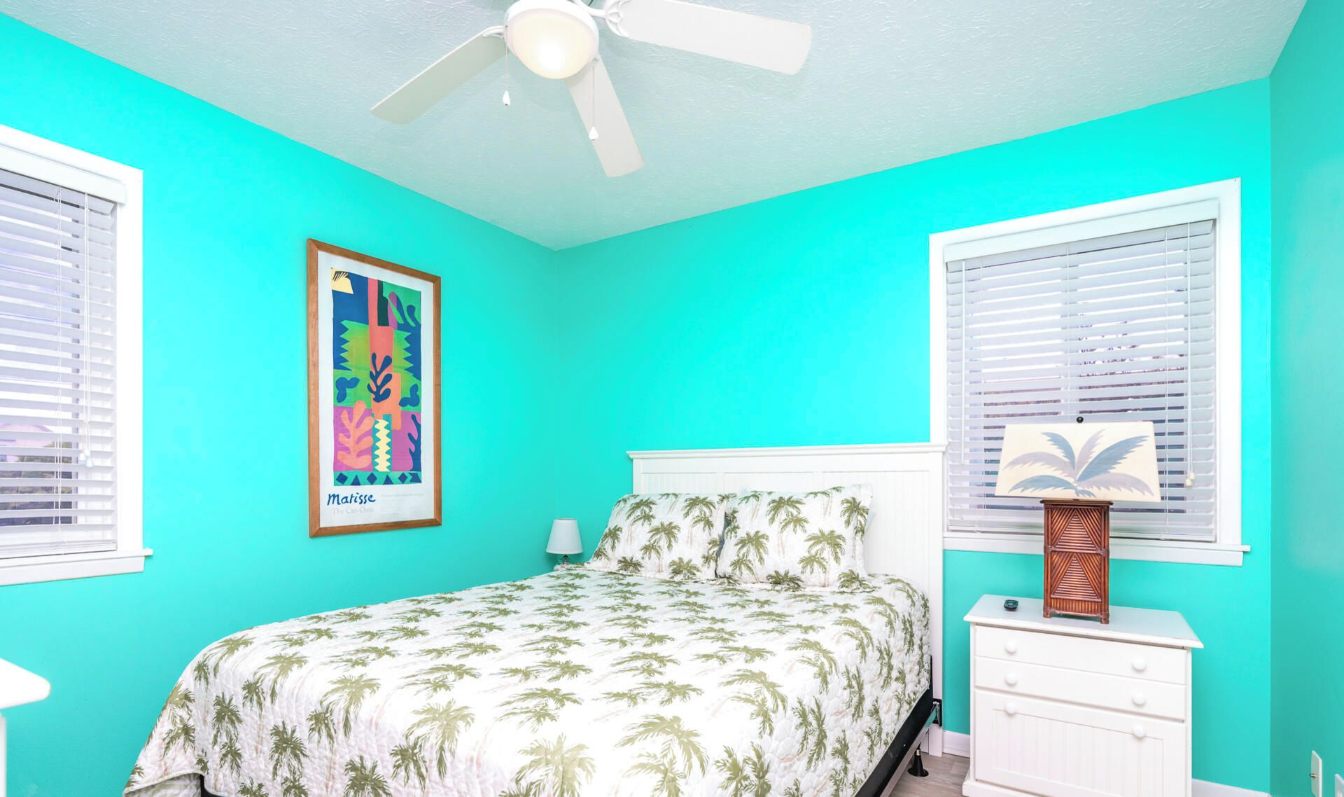 Quarto com cama, ventilador de teto, quadro na parede e mesa de cabeceira com abajur.
