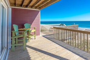 A wooden deck with a view of the ocean, a red building, and two green Adirondack chairs.