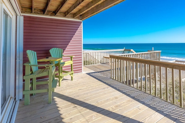 A wooden deck with a view of the ocean, a red building, and two green Adirondack chairs.