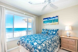 A bedroom with a bed, a nightstand, a lamp, a ceiling fan, a window with curtains, and a painting of a beach scene.
